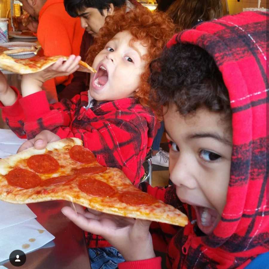 African American brothers, one with red curly hair, sit beside each other holding piece of pizza up to their mouth