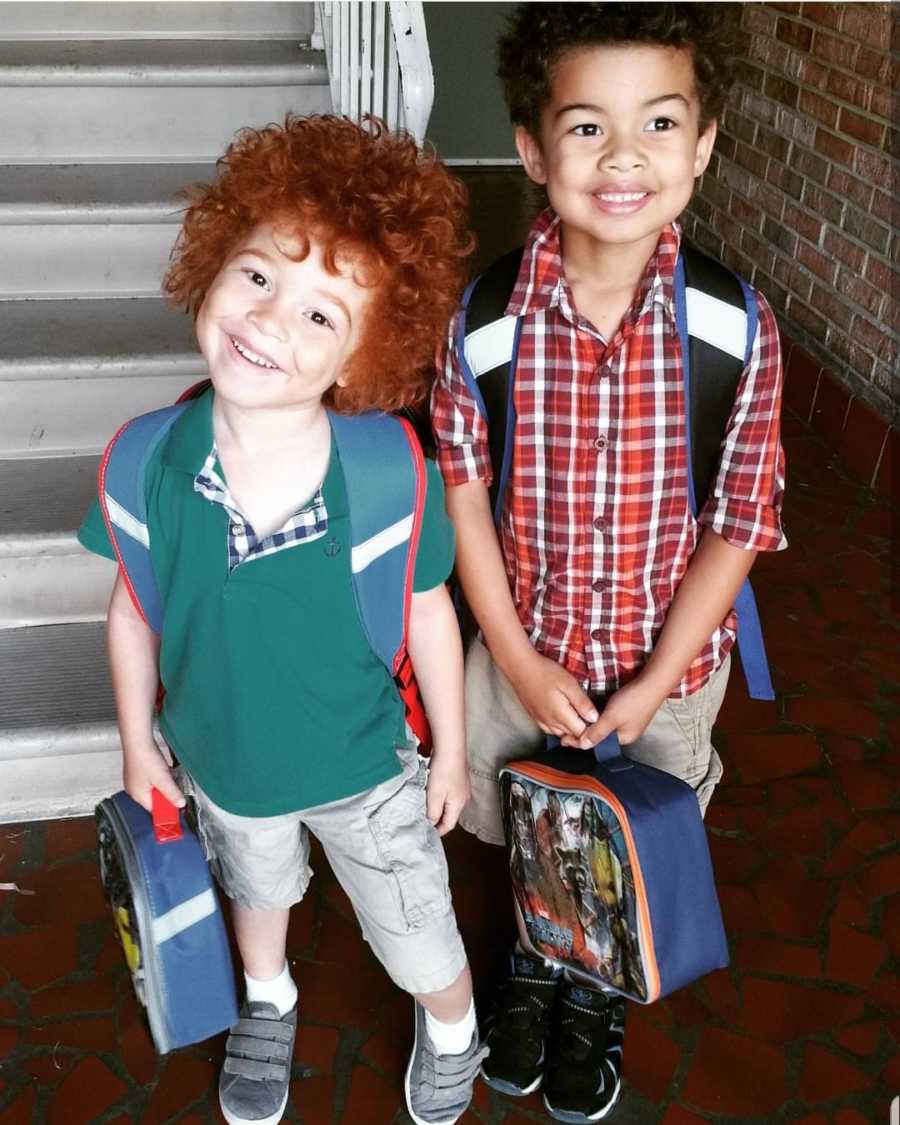 African American boy stands with brother with red curly hair as they both wear backpacks and hold lunchboxes 