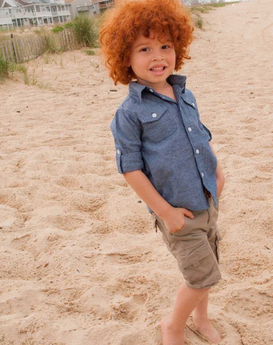 African American little boy with red curly hair stands on beach smiling with hands in his pockets