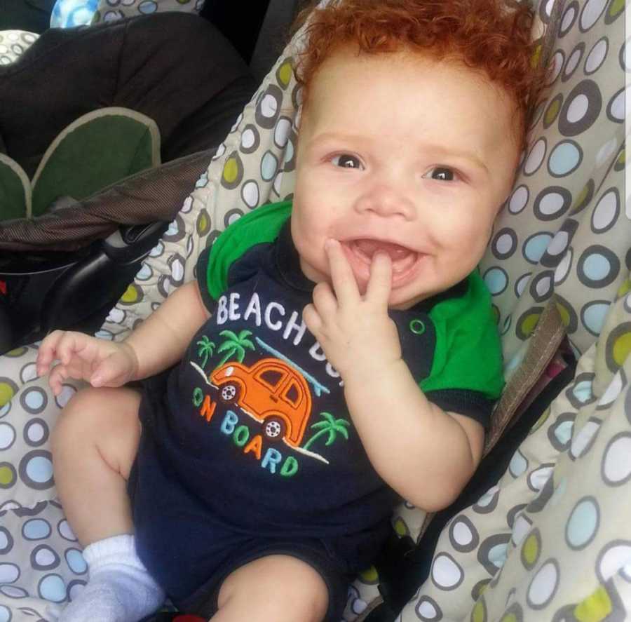 African American baby boy with red hair sits in car smiling with finger in mouth