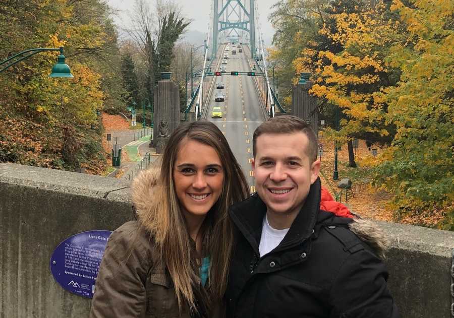 Couple who miscarried stands smiling in front of bridge with cars driving on it