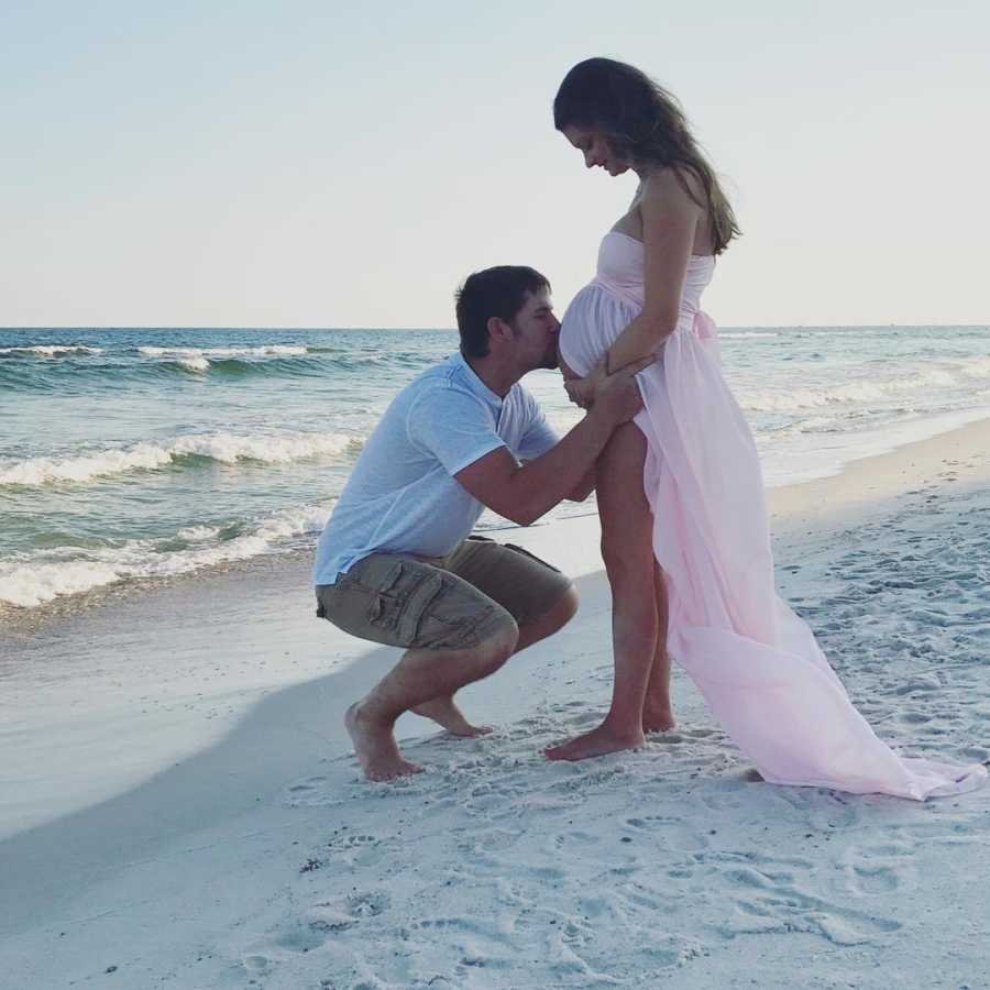 Pregnant woman stands on beach while husband crouches down kissing her stomach