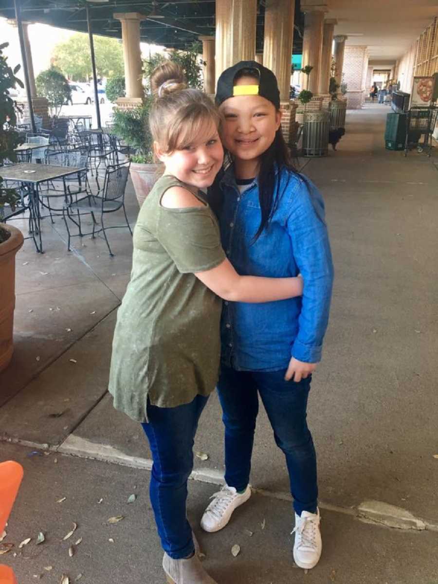 Parents biological daughter and adopted daughter hug near restaurant's outdoor tables