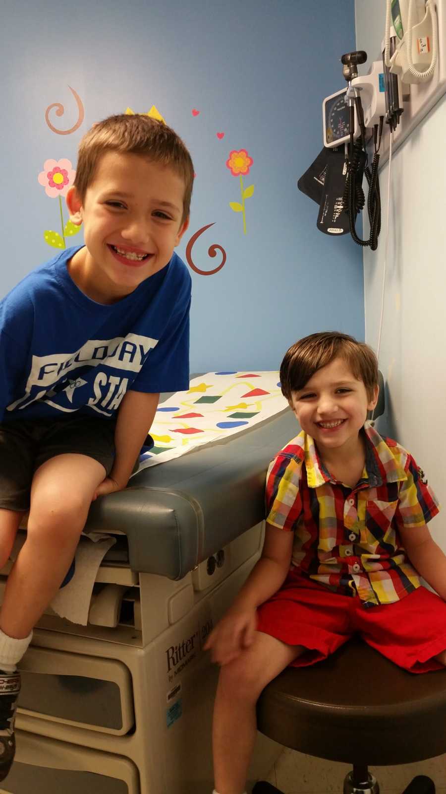 Young boy sits on bed in doctor's office beside younger brother who sits on doctor's stool