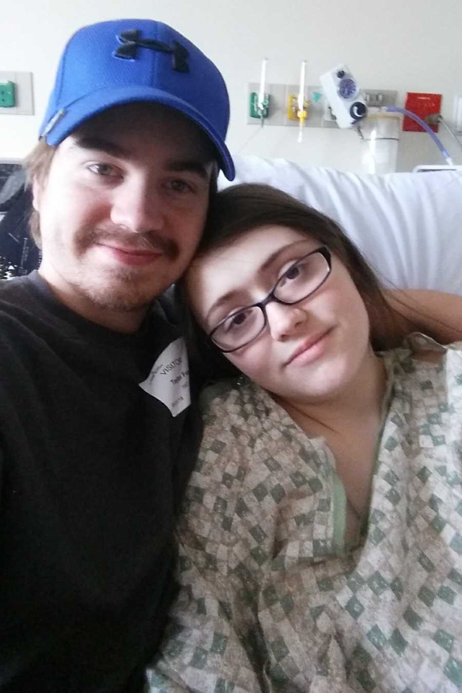 Husband stands smiling with arm around wife who sits in hospital bed in selfie