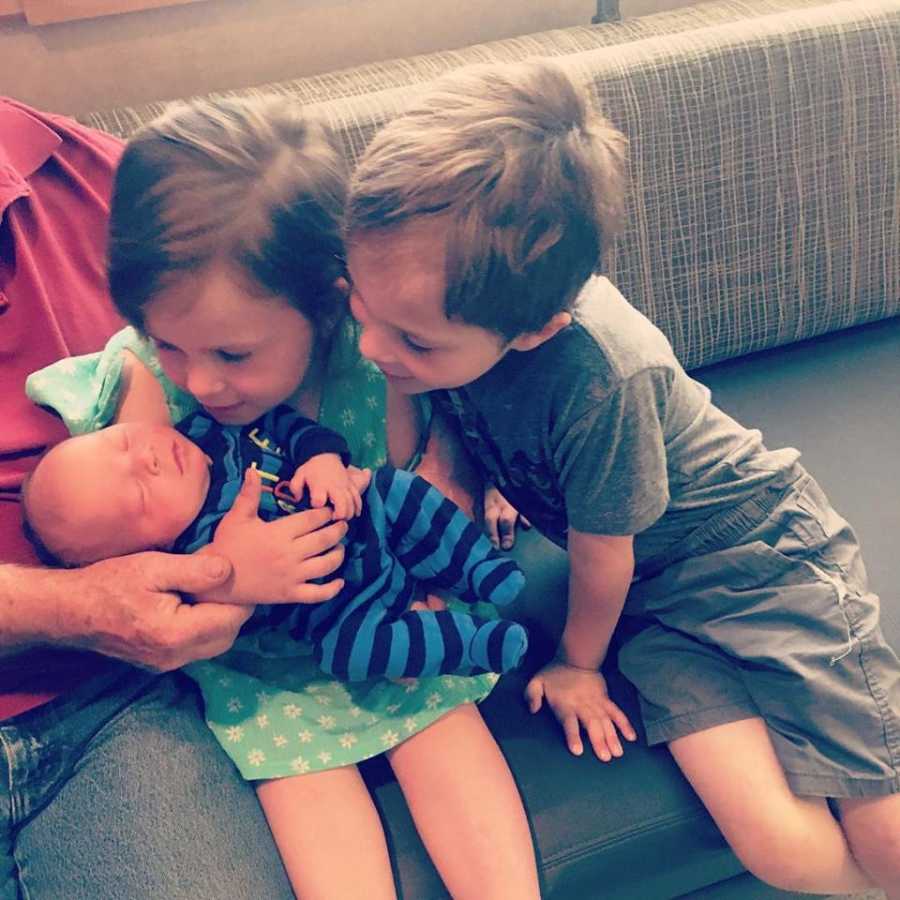 Older brother and sister sit on couch looking at newborn brother