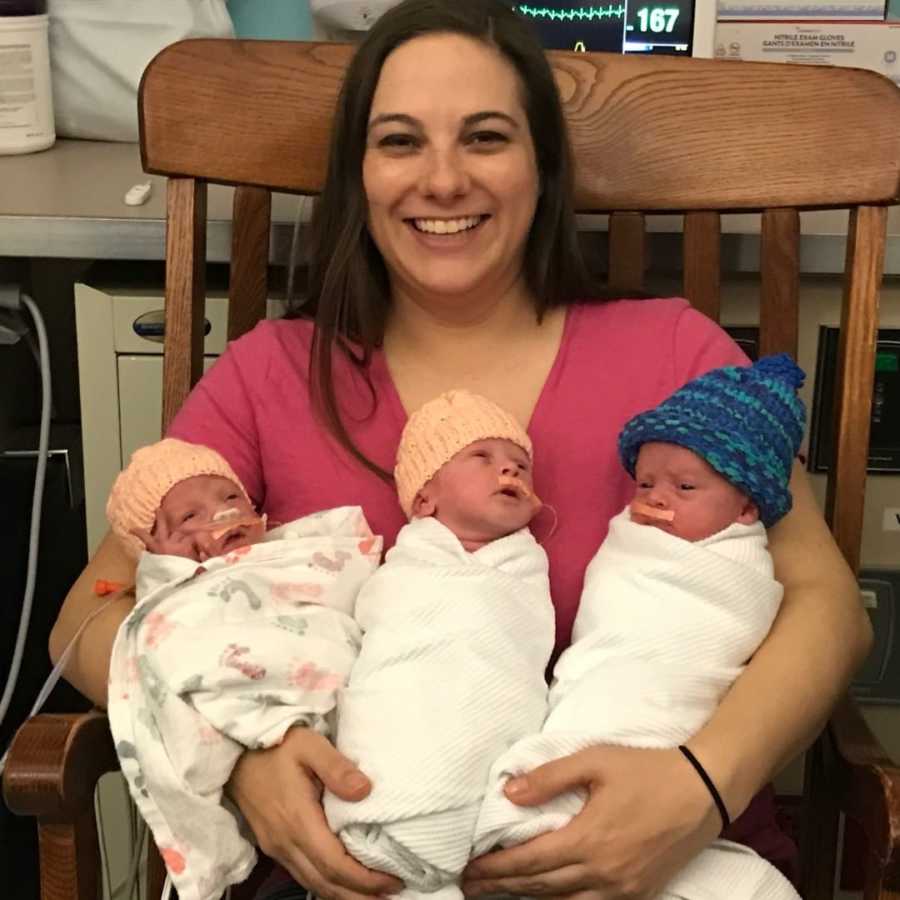 Mother smiles in rocking chair in NICU holding newborn triplets in her arms