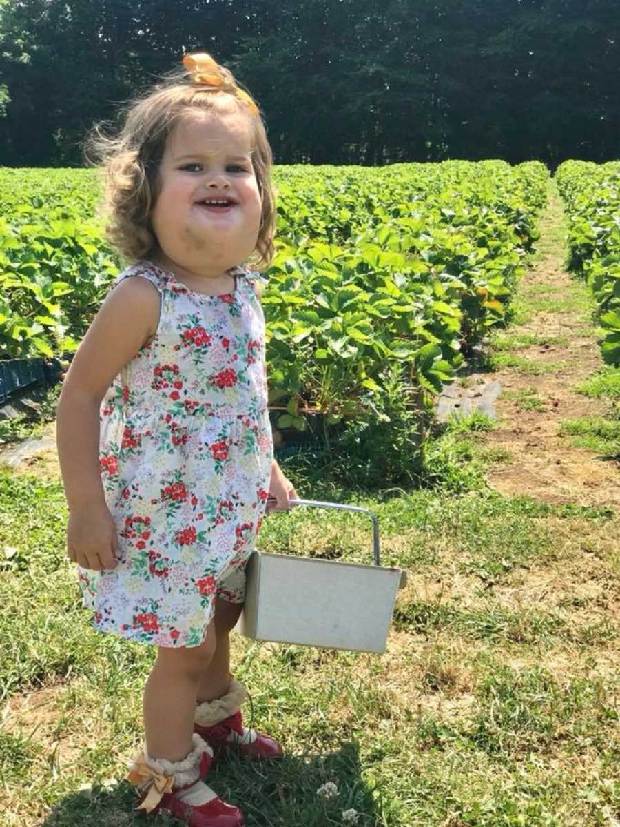 Little girl with cystic hygroma stands smiling holding bucket in berry field
