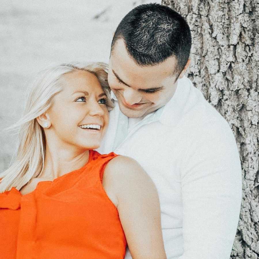 Husband leans against tree smiling while wife leans on him looking up at him