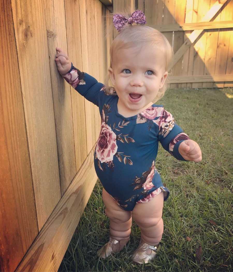 Little girl with mild form of dwarfism stands smiling in yard touching wood fence