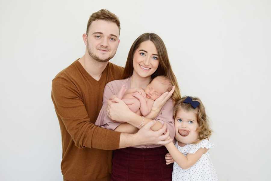 Husband and wife stand holding newborn beside first born with cystic hygroma