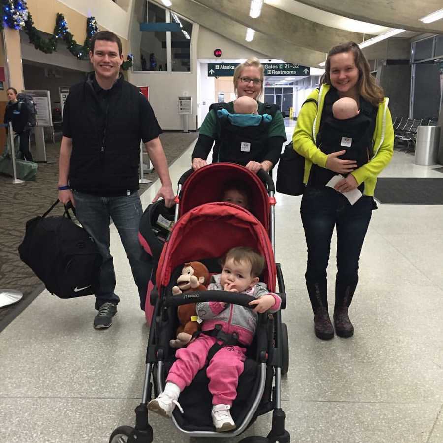 Wife and nanny stand in airport with baby swaddled to their chest while nanny also pushes stroller beside father who holds luggage
