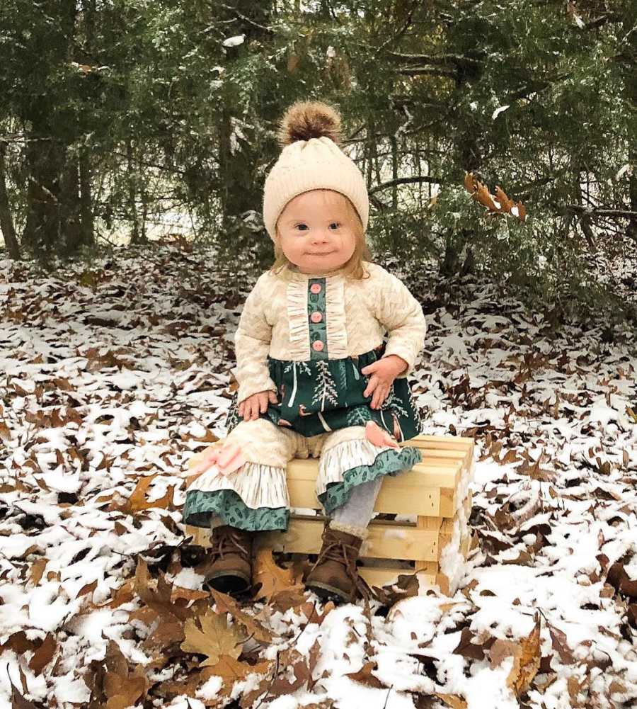Little girl with down syndrome sits on wooden crate outside with snowy leaves covering the ground