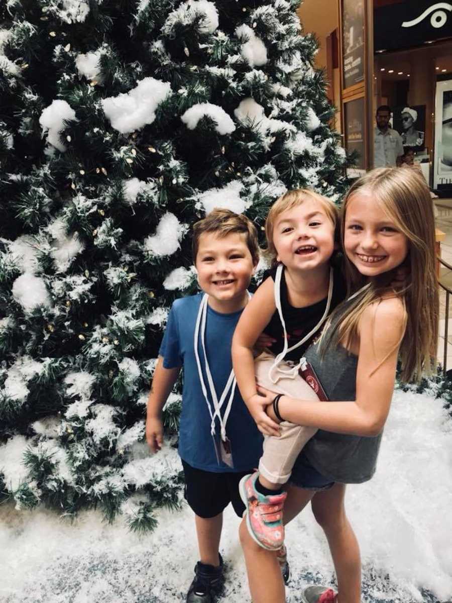 Oldest sibling holds youngest beside middle child as they stands smiling near Christmas tree covered in fake snow