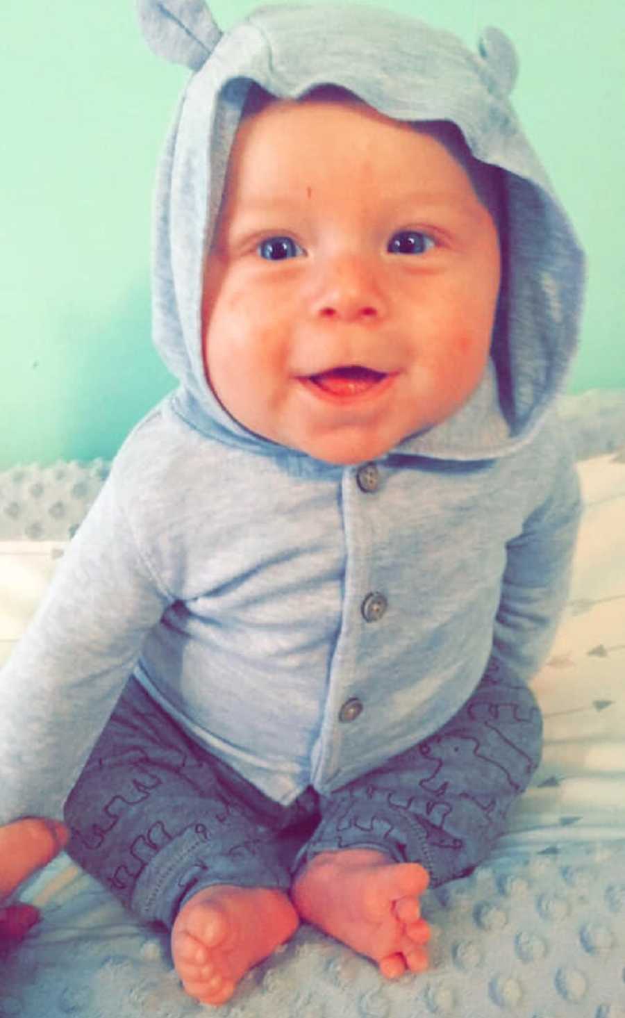 Baby with neuroblastoma smiles as he sits up wearing gray sweater that has hood with ears on it