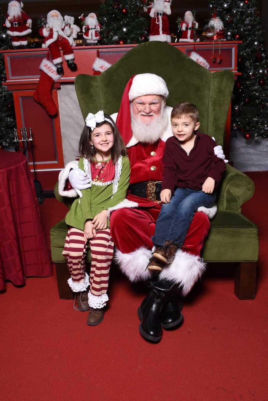 Santa sits in green chair with little girl and little boy with stomach tubes sitting on his lap