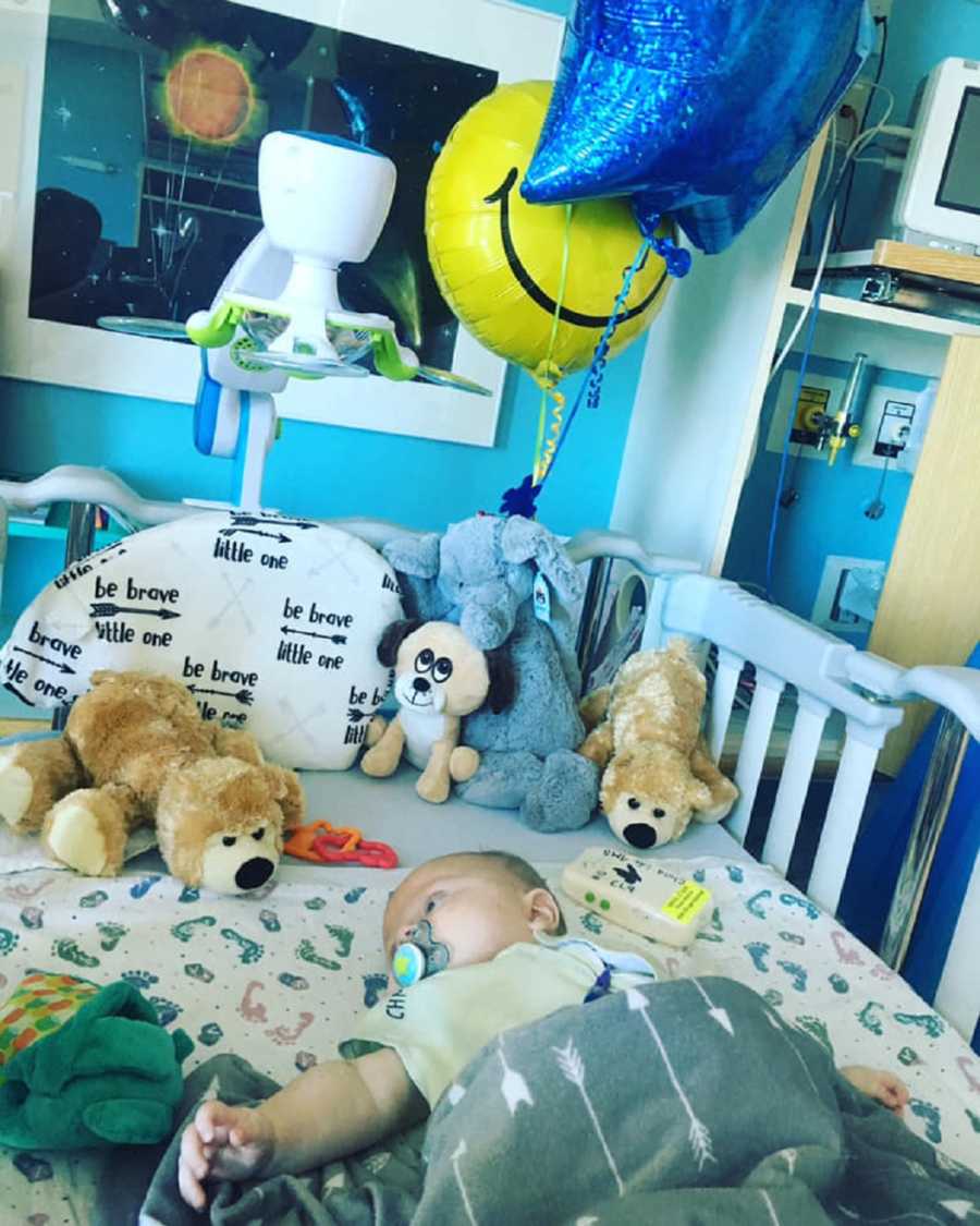 Baby with neuroblastoma lays in hospital crib surrounded by stuffed animals and balloons