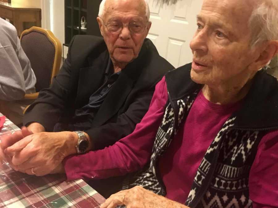 Elderly woman with dementia sits at table in home holding hands with husband 