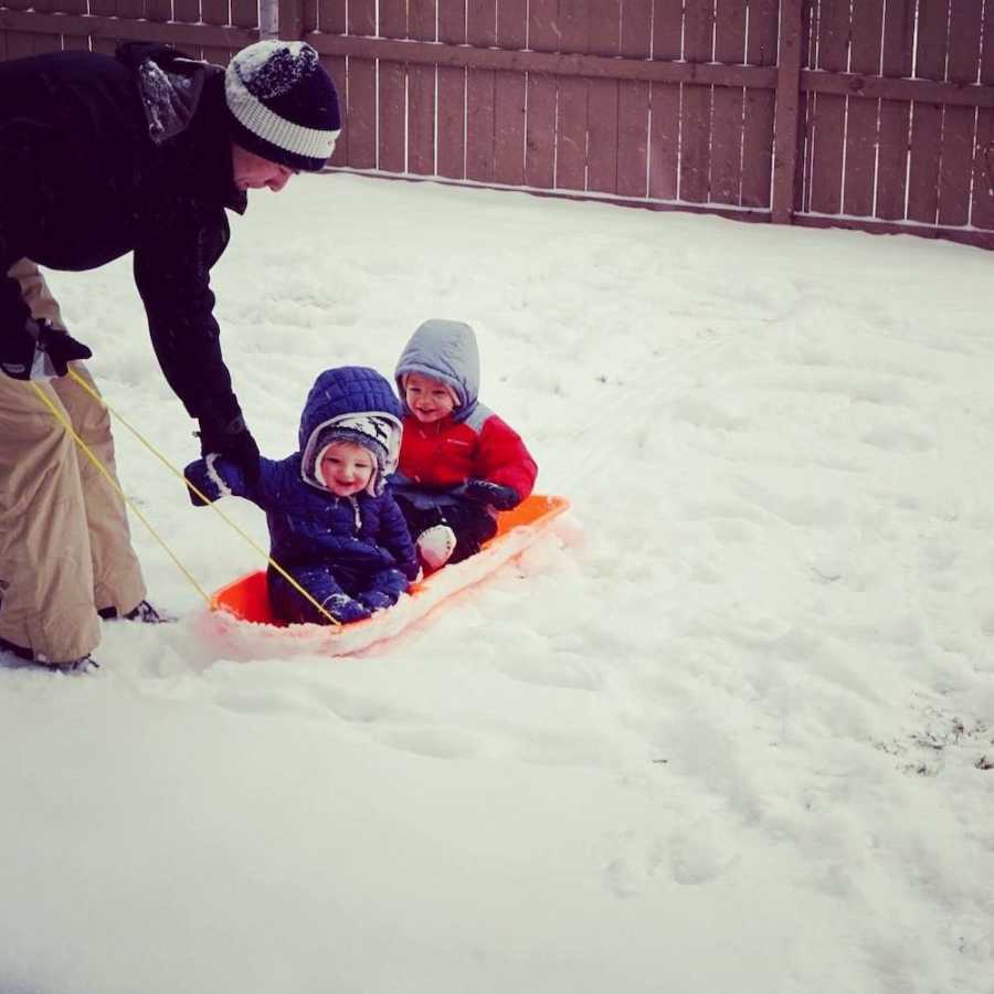 Two babies sit in orange sled in snowy yard while parent holds onto string of sled