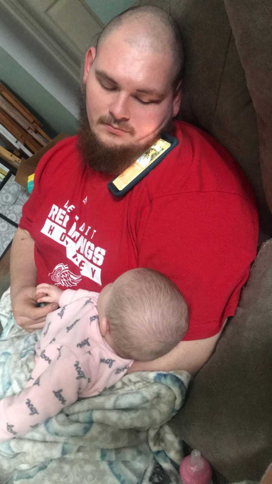 Father sits asleep in chair with phone on his shoulder and baby daughter in his lap