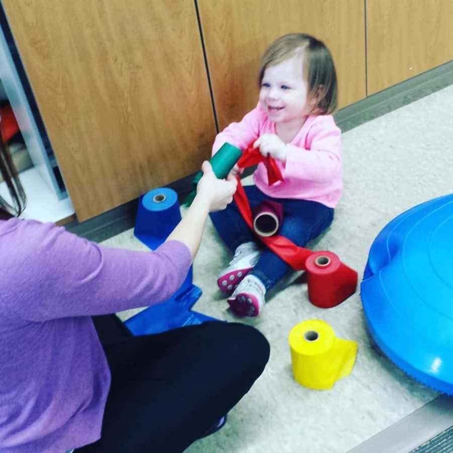 Little girl who had Hemorrhagic Stroke sits on floor holding red streamer as woman sits beside her holding out green streamer