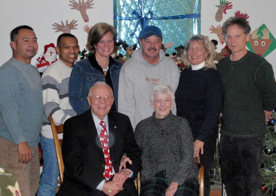 Elderly woman with dementia sits beside husband in chairs while family stands behind them