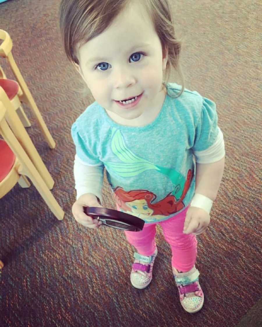 Little girl who had Hemorrhagic Stroke stands smiling wearing Ariel shirt