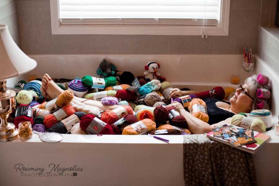 Woman with pink rollers in her hair lays in bathtub filled with bundles of yarn