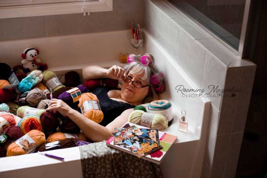 Woman with pink rollers in her hair holds hand to glasses as she lays in bathtub filled with bundles of yarn