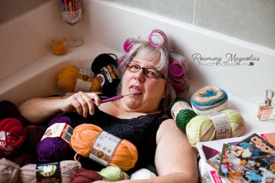 Woman with pink rollers in her hair holds knitting tool to mouth as she lays in bathtub filled with yarn