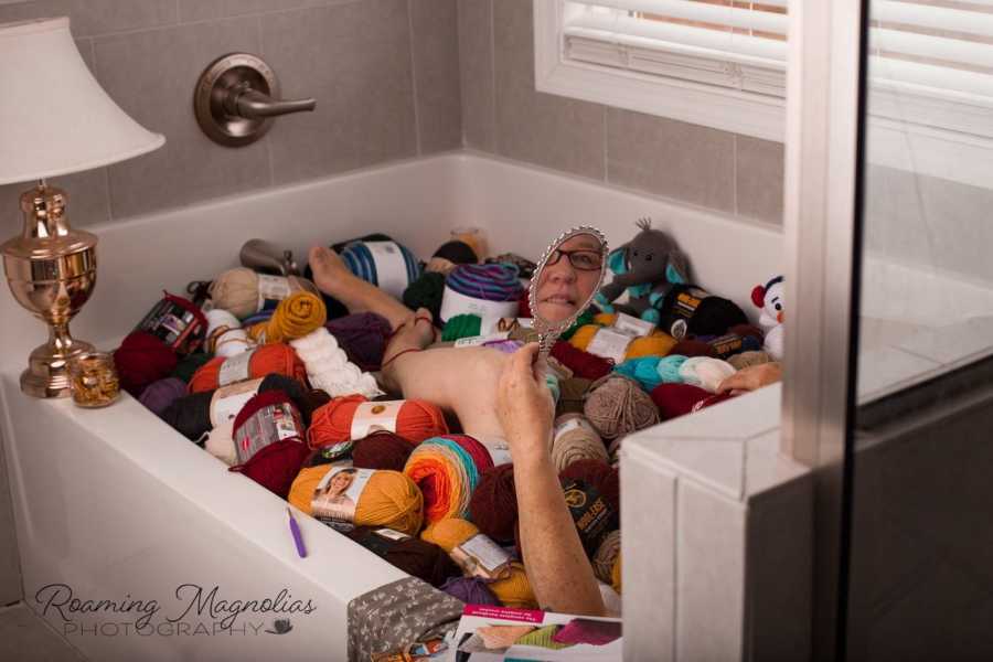 Woman holds up mirror to face as she lays in bath tub filled with yarn