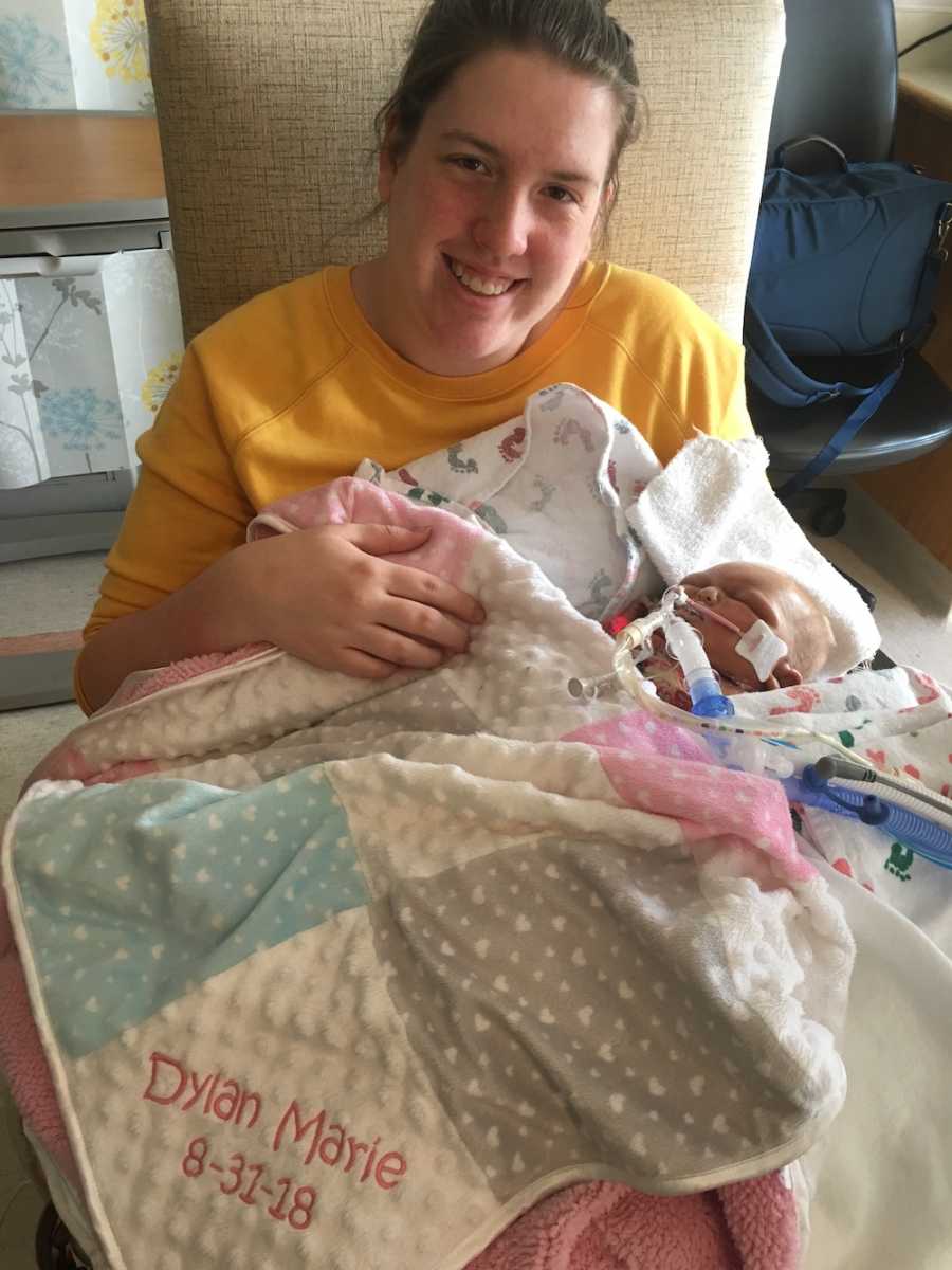 Woman smiles in NICU with newborn who went into renal failure laying on her lap with blanket covering her