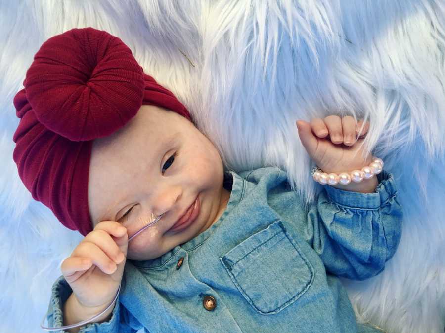 Baby with down syndrome lies on her back on white furry rug wearing denim top and maroon head wrap