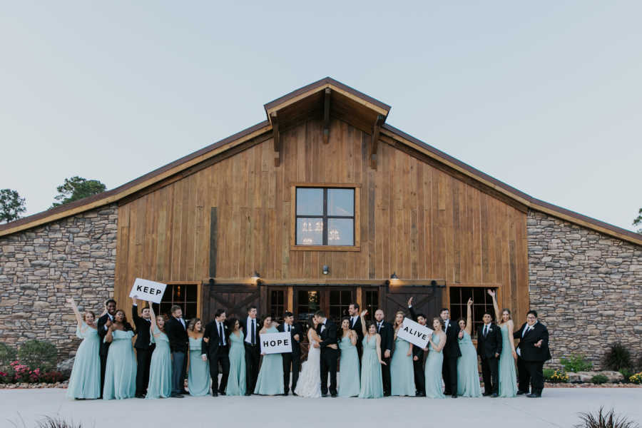 Wedding party stand on either side of bride and groom who are kissing holding signs that say, "keep hope alive"