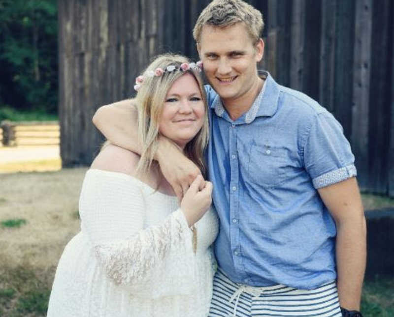 Husband and wife stand smiling with wooden barn in background 