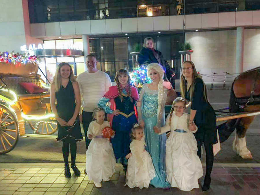 Husband and wife stand outside in front of horse and carriage with their two daughters, two foster daughters, and Elsa and Anna