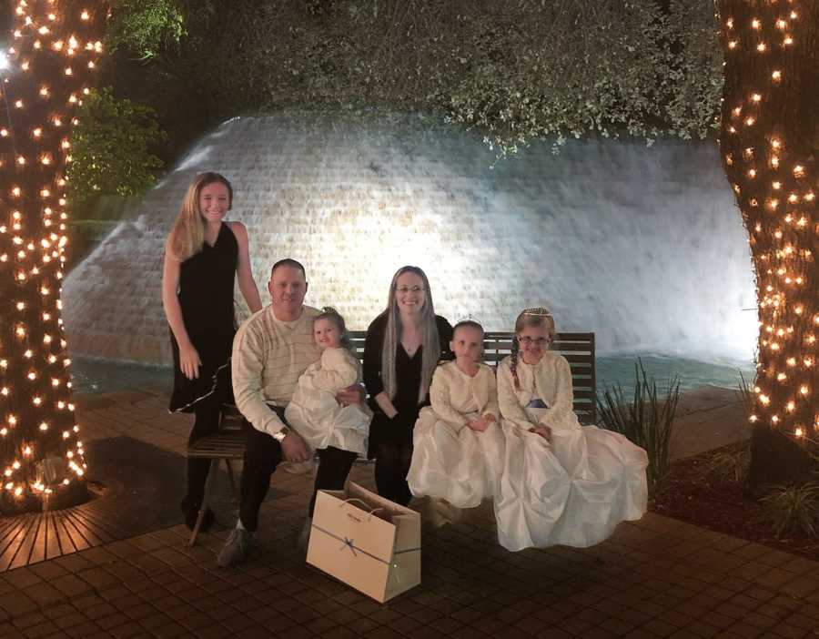 Husband and wife sit on bench outside beside their two daughter and two foster daughters between trees wrapped in lights