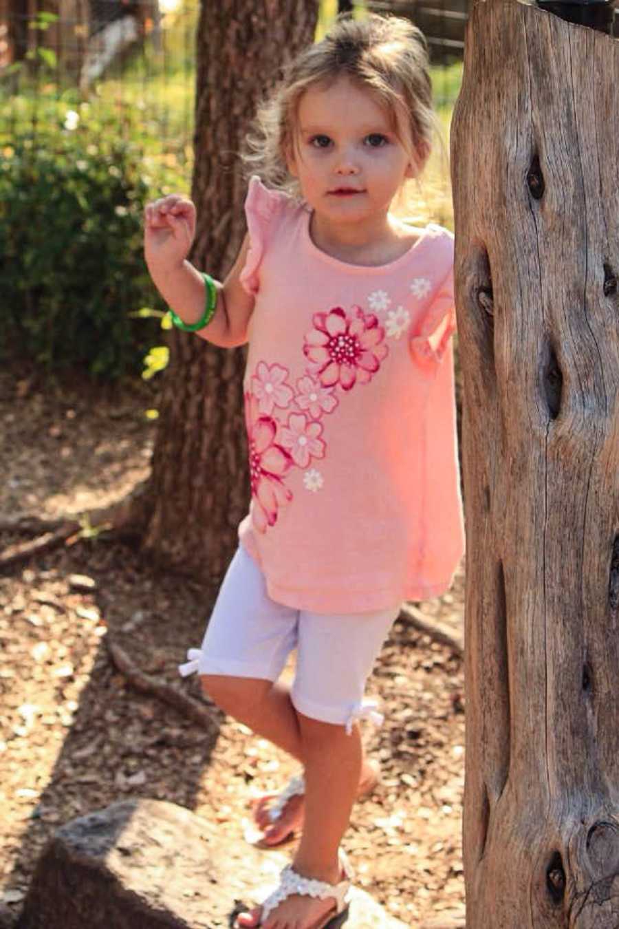 Little girl who was taken in by her hand stands leaning against tree wearing pink shirt with flowers and white shorts