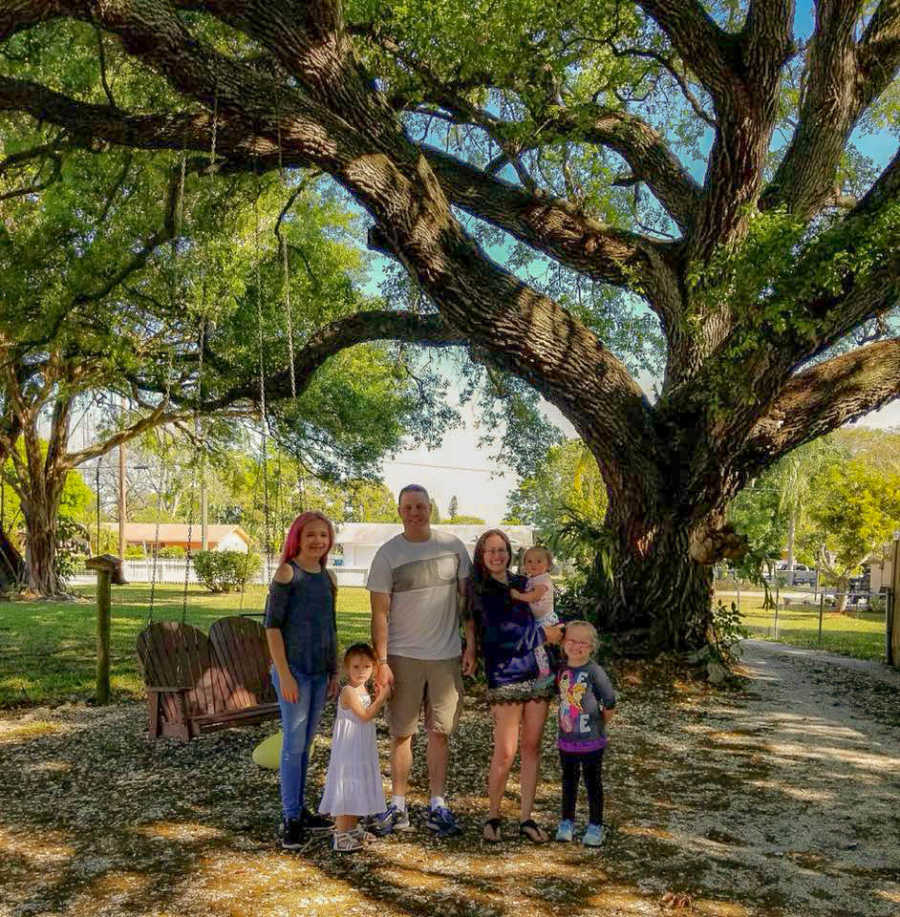 Husband and wife stand outside beside big tree with their two daughter and two foster daughters