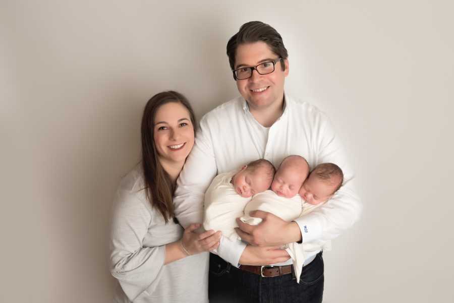 Smiling wife stands beside husband as he holds newborn triplets in his arms