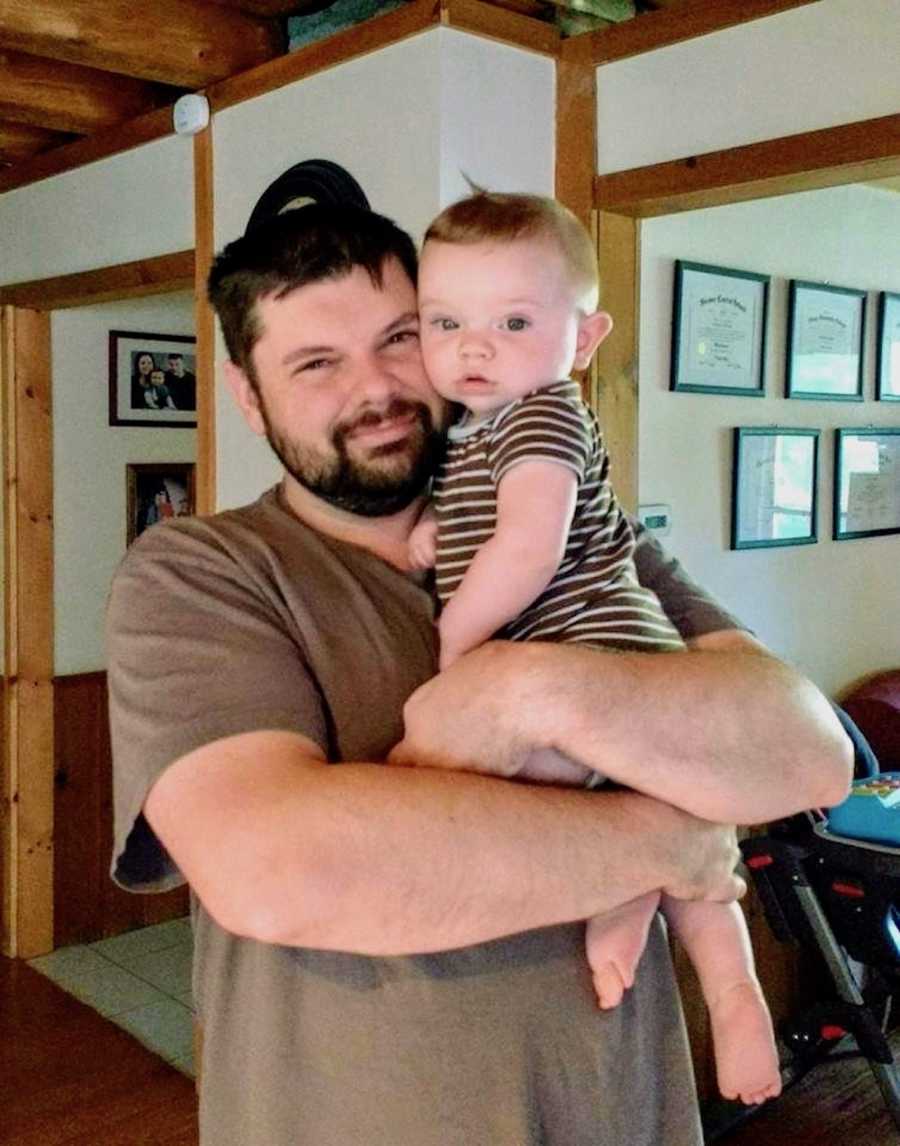 Husband, whose wife says he does it all, stands smiling in home holding baby son