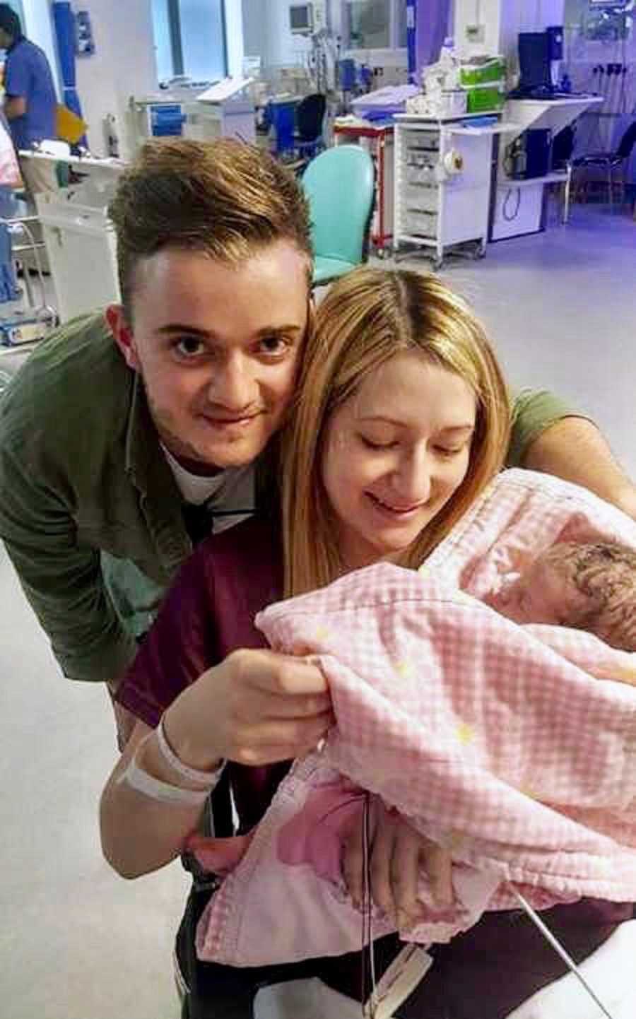 Husband smiles as he leans over beside wife who holds their newborn with lymphatic malformation in NICU