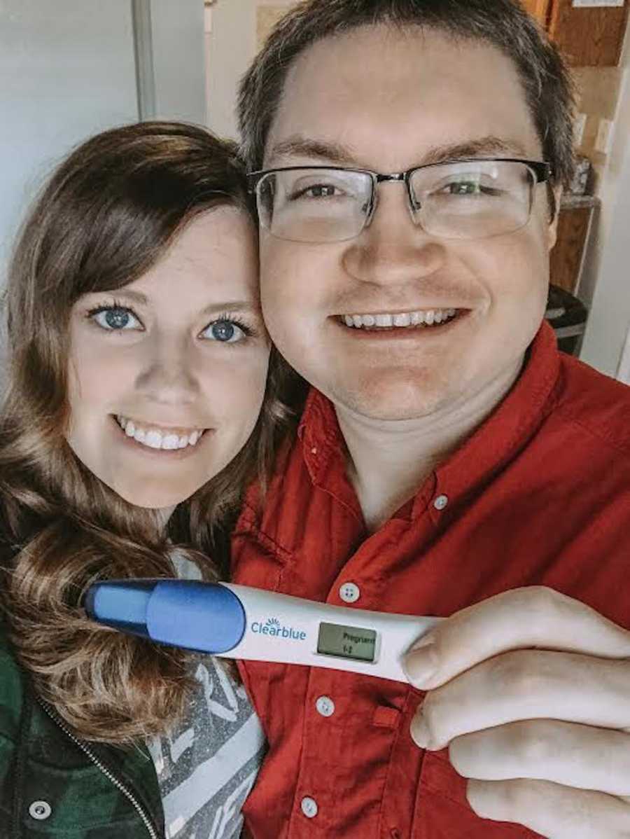 Husband and wife smile in selfie while husband holds up positive pregnancy test