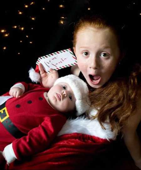 Little girl sits with mouth open while her baby brother sits on her lap in Santa suit