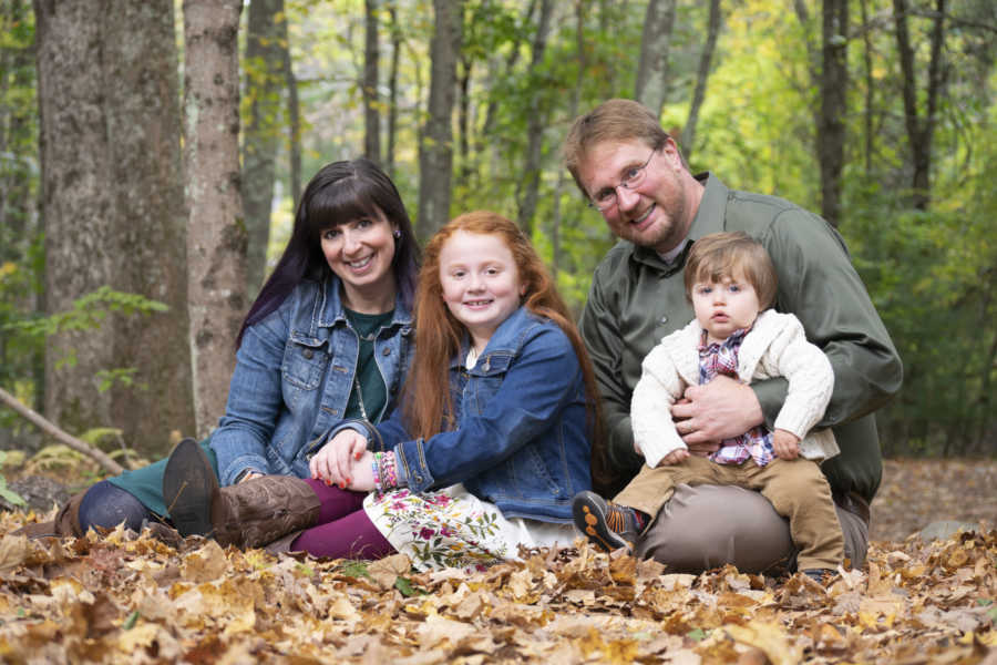 Husband and wife sit on pile of leaves outside with daughter and baby son