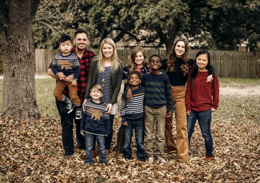 Husband and wife stands outside in pile of leaves with two biological kids and five adopted kids
