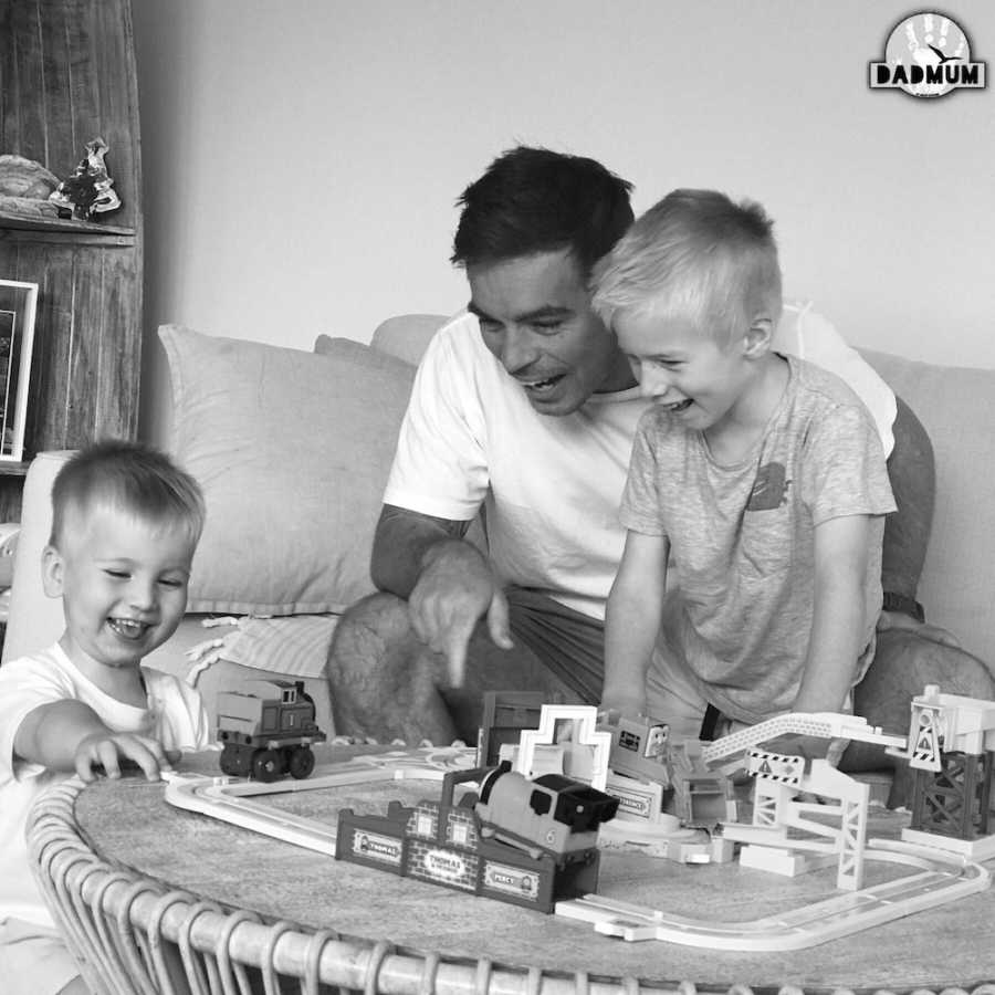 Father sits on couch watching his young sons play with toy train track on coffee table