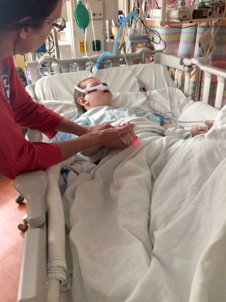 Young boy who had seizure lays in hospital bed while foster mom holds his hand