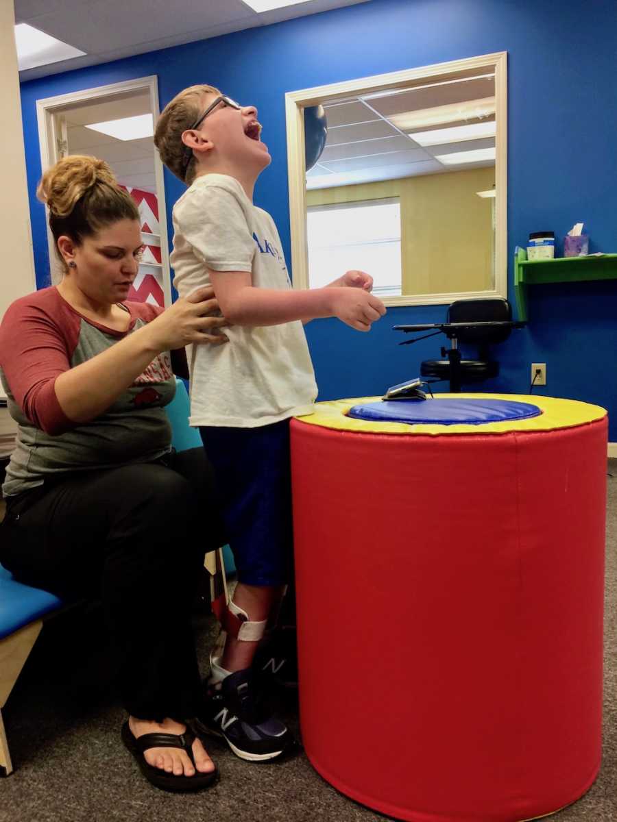 Young boy with shaken baby syndrome stands in therapy in front of foam circle while therapist holds him at his waist