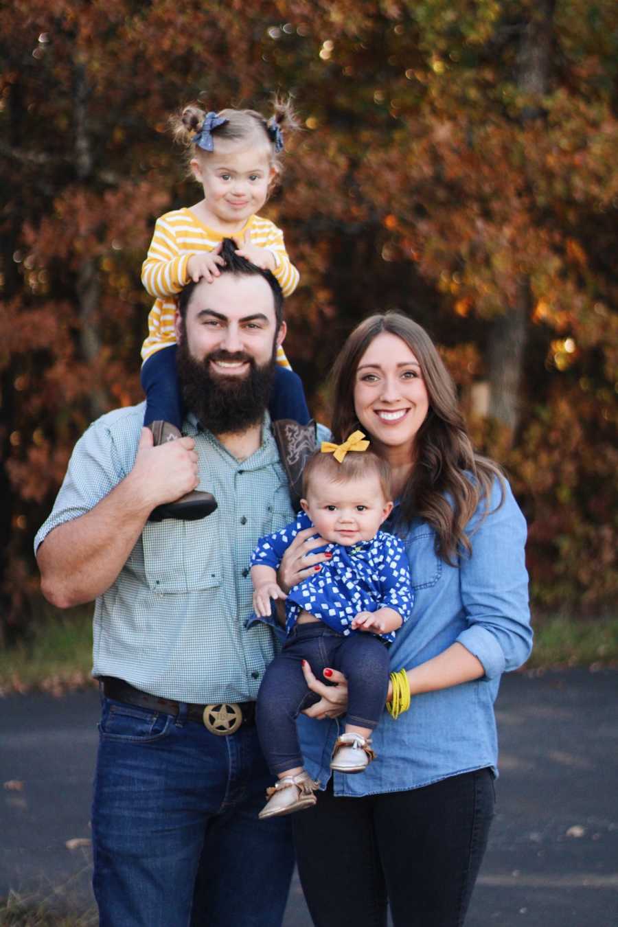 Mother smiles outside holding her baby daughter beside husband who has daughter with down syndrome on his shoulders